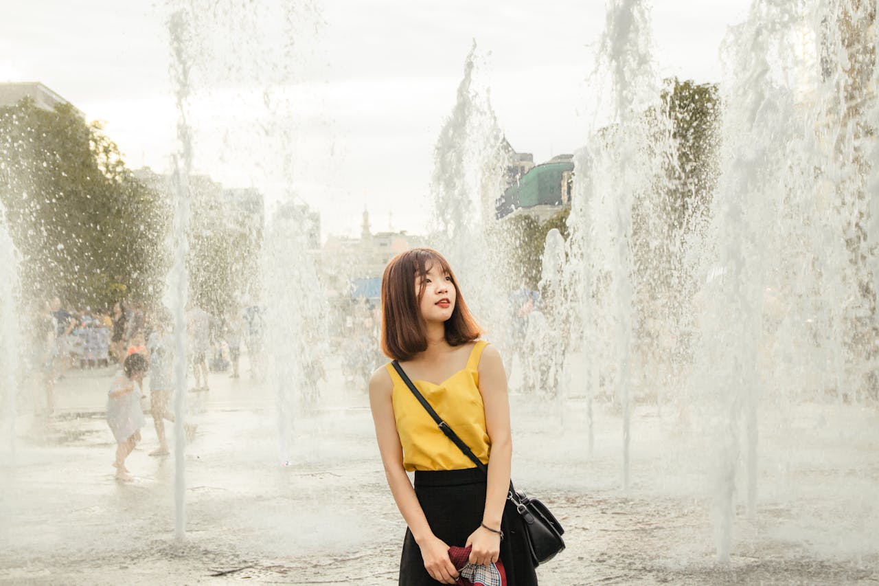 our-journey-01 A young woman in a yellow top stands near water fountains outdoors, showcasing a serene and fashionable scene.