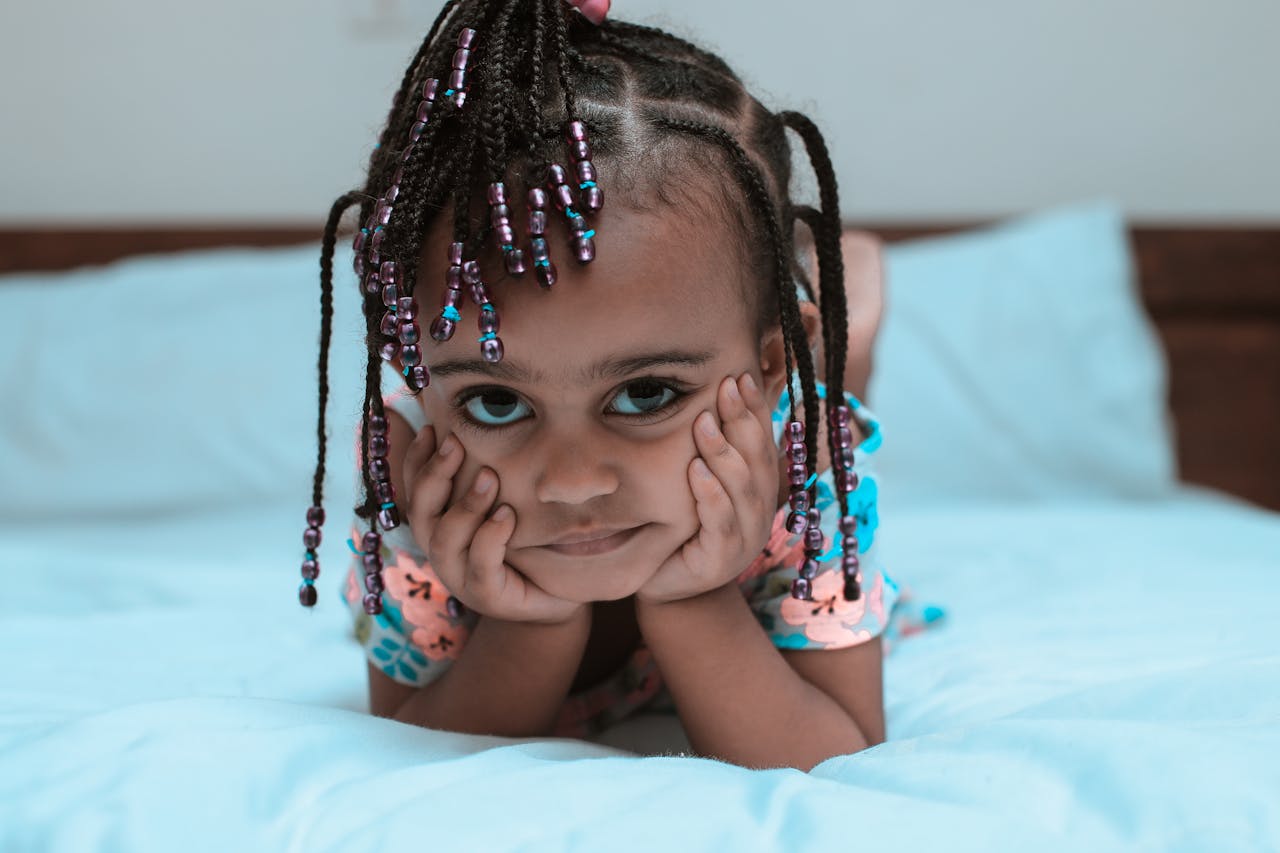 our-journey-03 Adorable child with braided hair and beads relaxing on a bed, showcasing innocence and style.