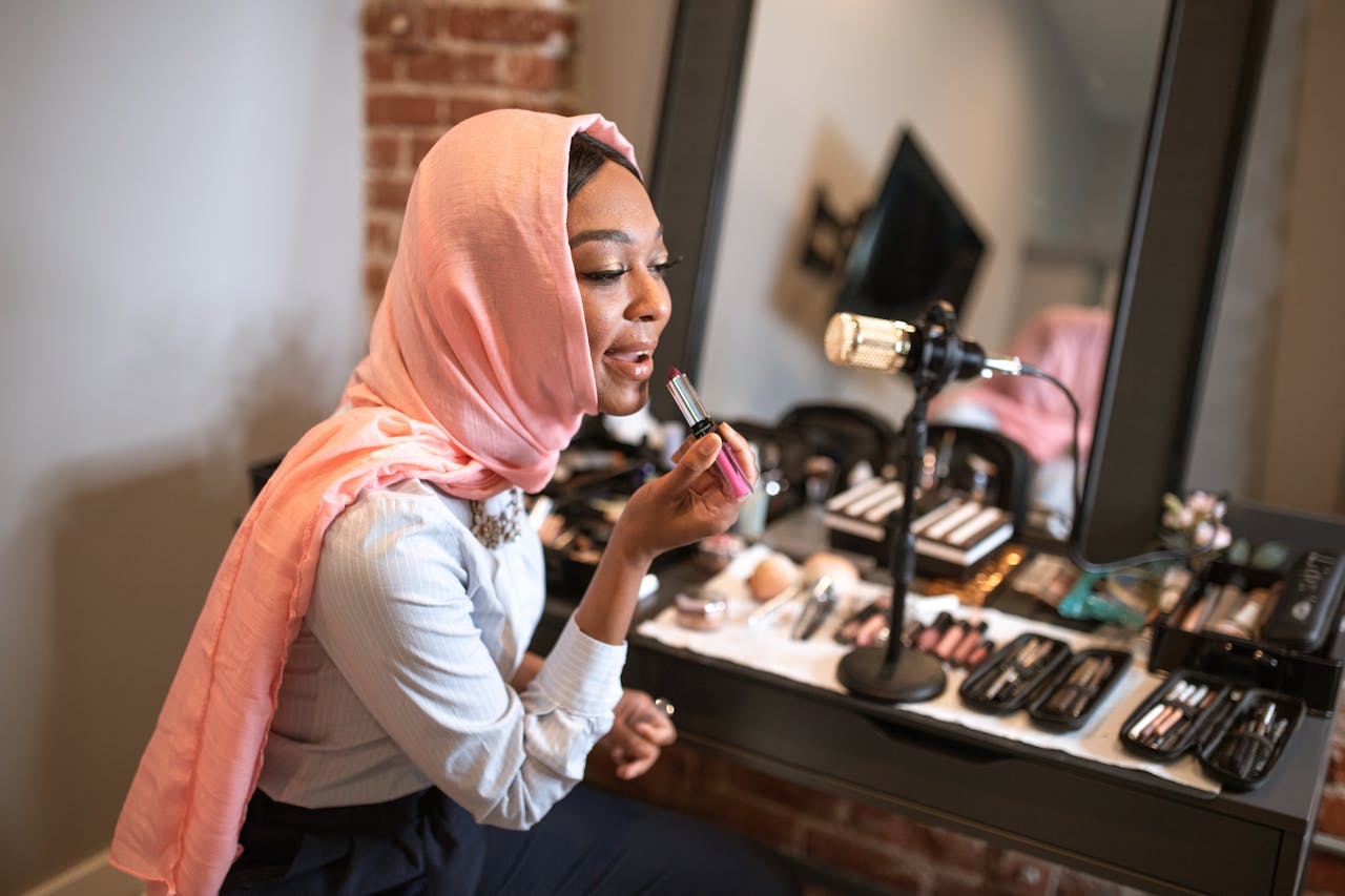 why-choose-us Muslim woman in pink hijab applying makeup while recording a beauty vlog indoors.